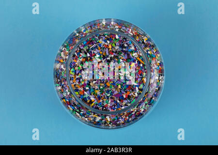 Crushed plastic pellets particles made of multicolored recyled plastics collected at sea in a glass jar, blue background ready to be used for new circ Stock Photo