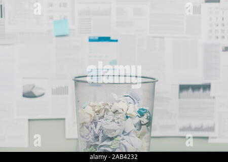 Trash can filled with crumpled paper and discarded paperwork, financial reports in the background Stock Photo
