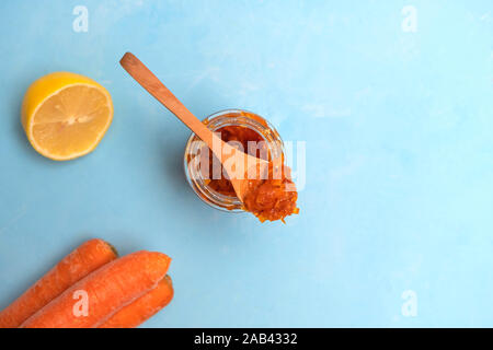 Homemade carrot jam in a glass jar with wooden spoon, fresh carrots and lemon on light blue background Stock Photo