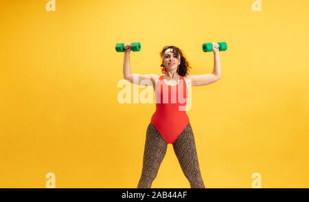 Young caucasian plus size female model's training on yellow background. Copyspace. Concept of sport, healthy lifestyle, body positive, fashion, style. Flexible woman with weights, looks stylish. Stock Photo