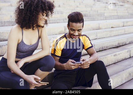 portrait of an athletic interracial couple having fun with mobile phone on the stairs during a rest in the workout, technology in sport and healthy li Stock Photo