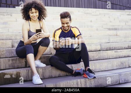 athletic interracial young couple having fun with mobile phone on the stairs during a rest in the workout, technology in sport and healthy lifestyle c Stock Photo
