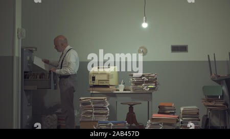 Corporate businessman searching for files in a rundown messy office with old computer and piles of paperwork Stock Photo