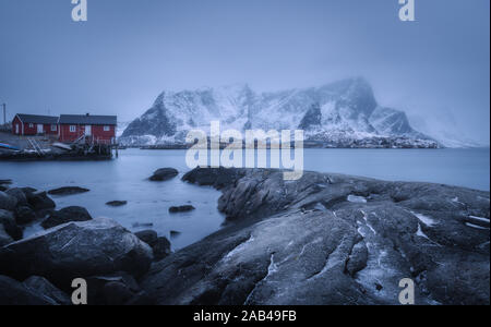 Beach with stones in blurred water, red rorbu and snowy mountains Stock Photo