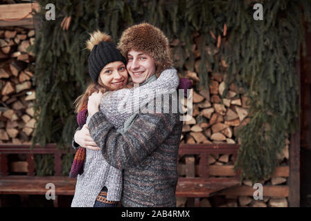 christmas and couple concept - smiling man and woman in hats and scarf hugging over wooden country house and snow background Stock Photo