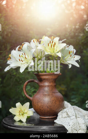 Still life with a cheerful bouquet of white lilies in a brown clay jug on a black table top in a Sunny Summer garden. Stock Photo