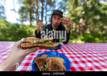 Selective focus of two adult people having a breakfast or lunch at a picnic table eating hot sandwiches while spending time in nature Stock Photo