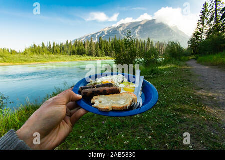 Selective focus of a persons hand holding up a blue plate full of sausages bread eggs and vegetables with amazing nature scenery in the background Stock Photo