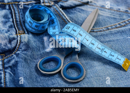 Measure tape wound around metal scissors on jeans. Tailors tools on denim fabric, selective focus. Jeans crotch, pocket and belt loops, close up. Making clothes and design concept. Stock Photo