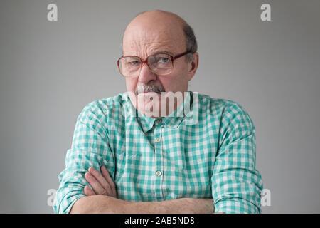 Angry senior man in glasses crossing arms looking annoyed at camera. He does not trust the news he is told. Studio shot Stock Photo