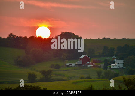 Bright orange sun setting over distant ridge, woodland in thriving farmland community, with farm, barns and crops in fields, Browntown, Wisconsin, USA Stock Photo