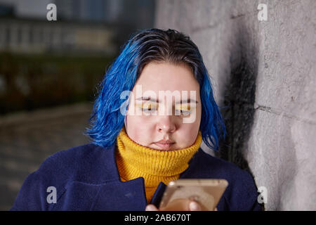 Attractive young woman with blue hair about 21 years old is standing outside and looking on the screen of the mobile phone in her hands. Caucasian ove Stock Photo