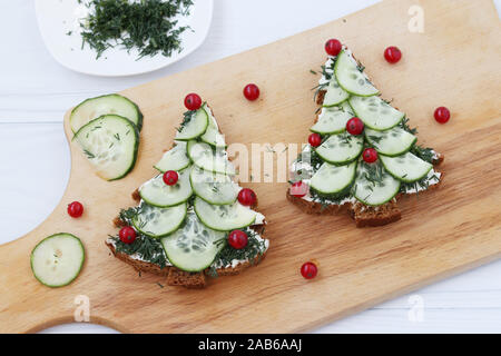 Sandwiches made of black bread, cheese and cucumber in the form of a Christmas tree decorated with berries, are placed on a wooden board on a white Stock Photo
