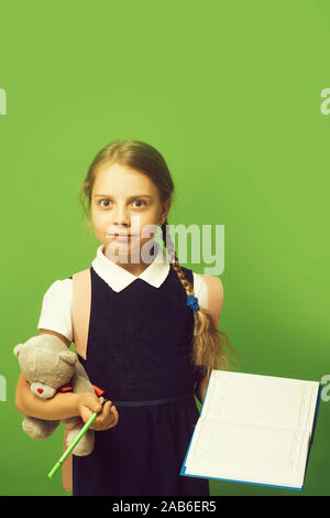 Pupil holds blue book, marker and teddy bear. Kid in school uniform isolated on green background. Girl with braids and surprised face expression. Study and back to school concept. Stock Photo