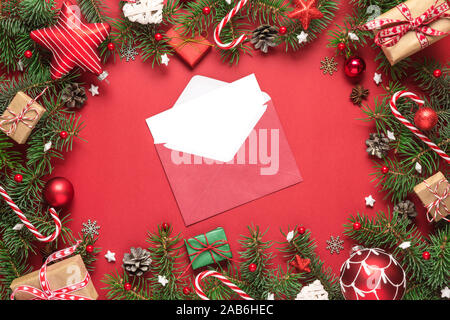 Christmas greeting card in frame made of fir tree branches, gift boxes, candy, holiday decorations on red background. mock up. flat lay. top view with Stock Photo
