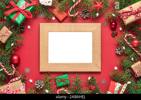 Christmas photo card in frame made of fir tree branches, gift boxes, candy, holiday decorations on red background. mock up. flat lay. top view with co Stock Photo