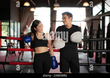 Asian boxer sports girl and trainer looking  smile while leaned on black red ropes on boxing ring, and have a rest after hard training in black loft g Stock Photo