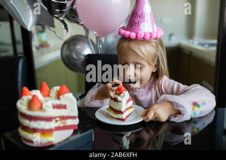 Indoor shot of pretty joyful little girl with blonde hair blowing out the candle, celebrate 6 years old birthday, wear fashionable dress, have excited Stock Photo