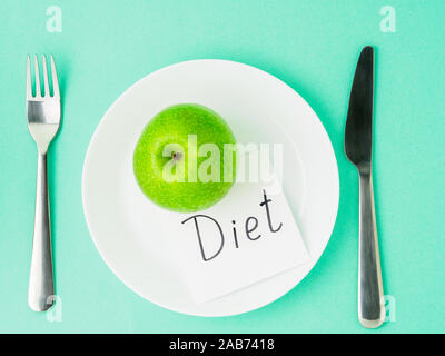Ripe raw juicy green Apple, slip of paper with inscription diet on a white plate, fork, knife on a bright turquoise blue colour table, top view. Diet Stock Photo