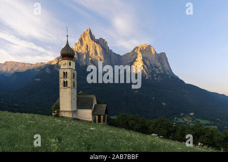 Sunrise landscapes of Church St. Valentin on grassy hilltop with view of rugged peaks of Mountain Schlern with alpenglow in background in the valley o Stock Photo