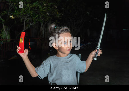A cute boy with a spiky hairstyle playing with a plastic sword in the dark. Stock Photo