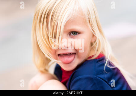 A cute, pretty little blonde haired girl wearing a football shirt being cheeky, smiling and having lot's of fun outdoors in the UK countryside Stock Photo