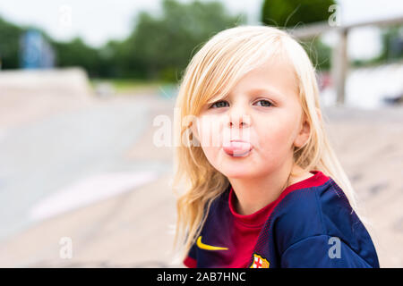 A cute, pretty little blonde haired girl wearing a football shirt being cheeky, smiling and having lot's of fun outdoors in the UK countryside Stock Photo