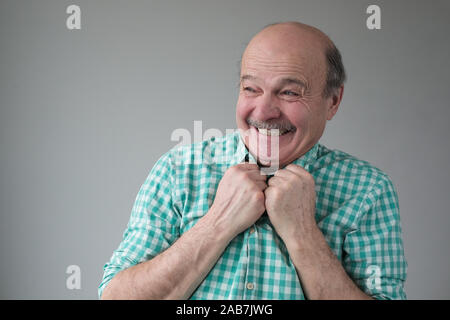 Surprised senior man in denim shirt looking aside keeping hands near chest while standing against grey background Stock Photo