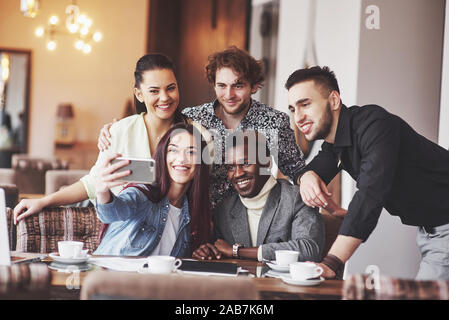Friends having fun at restaurant. Three boys and two girls making selfie and laughing. On foreground boy holding smart phone. All wear casual clothes Stock Photo