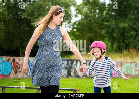 A young girl wearing a pink helmet holds her mothers hand as she learns to roller skate at the skatepark, fit, young and healthy exercise Stock Photo