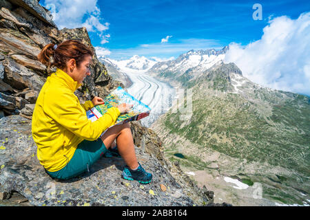 Woman looking at map sitting on rocks at Eggishorn viewpoint above Aletsch Glacier, Bernese Alps, canton of Valais, Switzerland, Europe Stock Photo