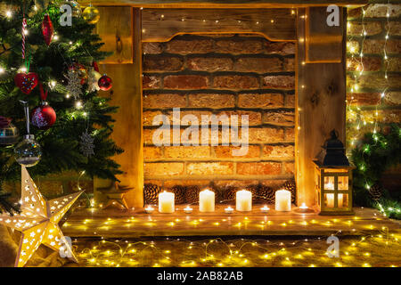 Beautiful decorated fireplace, wooden mantelpiece with fairy lights, handmade ornaments, candles and lantern, Christmas tree to the side, selective focus Stock Photo