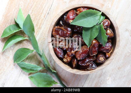 Wooden bowl full of dried dates with herb leaves. Sweet fruit - fruits of the date palm (Phoenix dactylifera). Brown bowl on teak table. Stock Photo