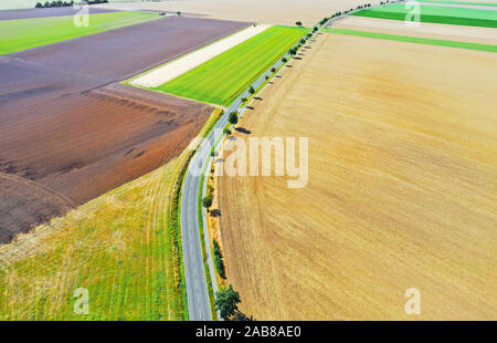 Aerial view of a grey asphalted narrow country road, which runs arcuately through a landscape with fields and meadows. Stock Photo