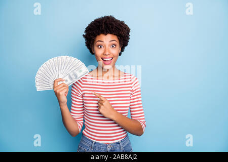 Photo of pretty sweet stunned girlfriend with astonishment on face smiling toothily pointing at fan of money she holds in striped shirt jeans denim Stock Photo