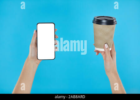 Woman hands holding coffee paper cup and smartphone on a blue background. Smartphone mockup with white screen Stock Photo
