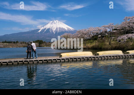 Japan, Fujinomiya, Shizuoka Prefecture: Mount Fuji with its snow-covered summit viewed from Lake Tanuki. Couple of Japanese tourists on a pontoon look Stock Photo