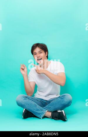 Studio shot of handsome stylish caucasian man with bristle sitting with crossed legs on floor while pointing left and smiling broadly, expressing posi Stock Photo