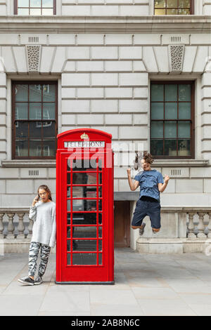 Girl is leaning against the phone booth with a smartphone and from the other side a boy leaps with joy. Stock Photo