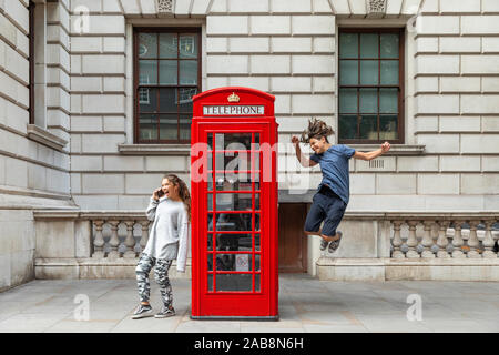 Girl is leaning against the phone booth with a smartphone and from the other side a boy leaps with joy. Stock Photo