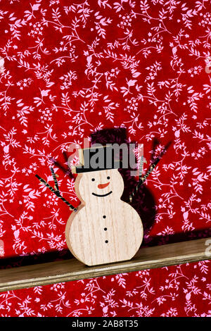 a funny wooden snowman, with a red nose and wearing a hat, on a wooden shelf against a floral-patterned wallpapered wall Stock Photo