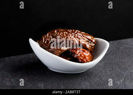 some pieces of sun-dried tomatoes in a white ceramic bowl on a dark gray stone surface, against a black background Stock Photo