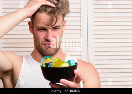 Fitness lifestyle and regime idea. Man with unshaven face holds bowl with measuring tape. Weight management and sportive diet concept. Guy with thoughtful face expression on beige jalousie background Stock Photo