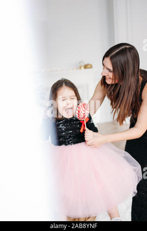Young mother and daughter with red candy in evening dresses on background of Christmas tree Stock Photo