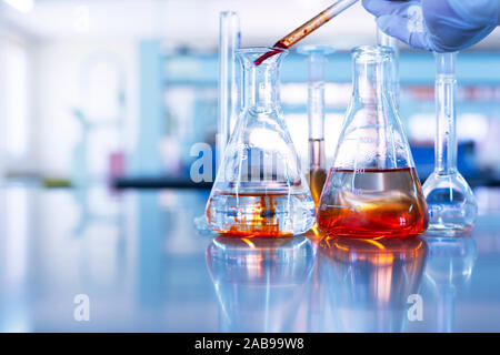 hand of scientist drop orange solution by pipette in glass flask in chemistry science laboratory blue background Stock Photo