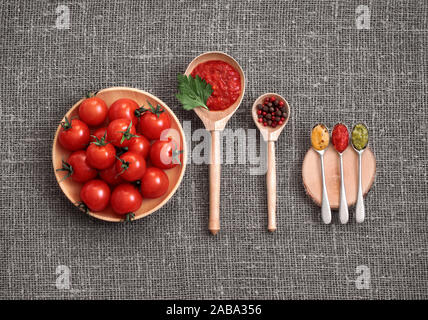 Cherry tomatoes in a wooden plate on a gray background. Adjika sauce, red and black pepper, parsley, mustard and pesto in spoons on burlap. Top view, Stock Photo