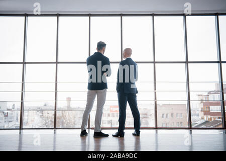 Two businessmen deep in discussion together while standing in an office boardroom with windows overlooking the city Stock Photo