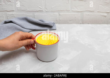 Woman hand holding cup of indian drink turmeric golden milk on concrete kitchen table. Stock Photo