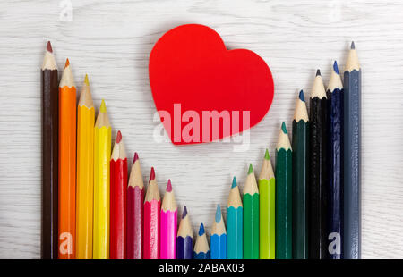 Series of colored pencils on a white wood background with a red heart-shaped box in the middle Stock Photo