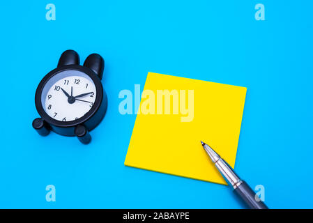 Flat lay of an alarm clock, pen and yellow paper to write any text, isolated on plain blue background. Stock Photo